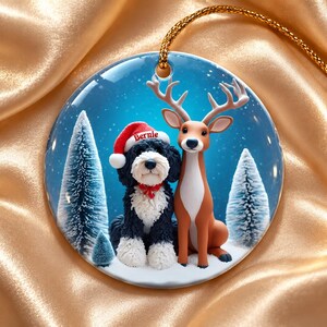 May include: A round ceramic ornament featuring a black and white dog wearing a red Santa hat with the name Bernie printed on it, sitting next to a brown deer. The background shows snowy pine trees under a blue sky with falling snow.