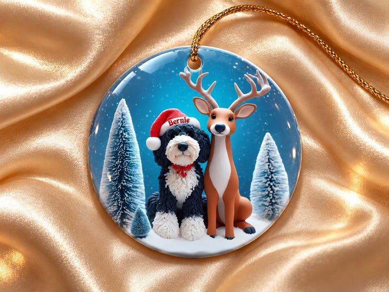 May include: A round ceramic ornament featuring a black and white dog wearing a red Santa hat with the name Bernie printed on it, sitting next to a brown deer. The background shows snowy pine trees under a blue sky with falling snow.