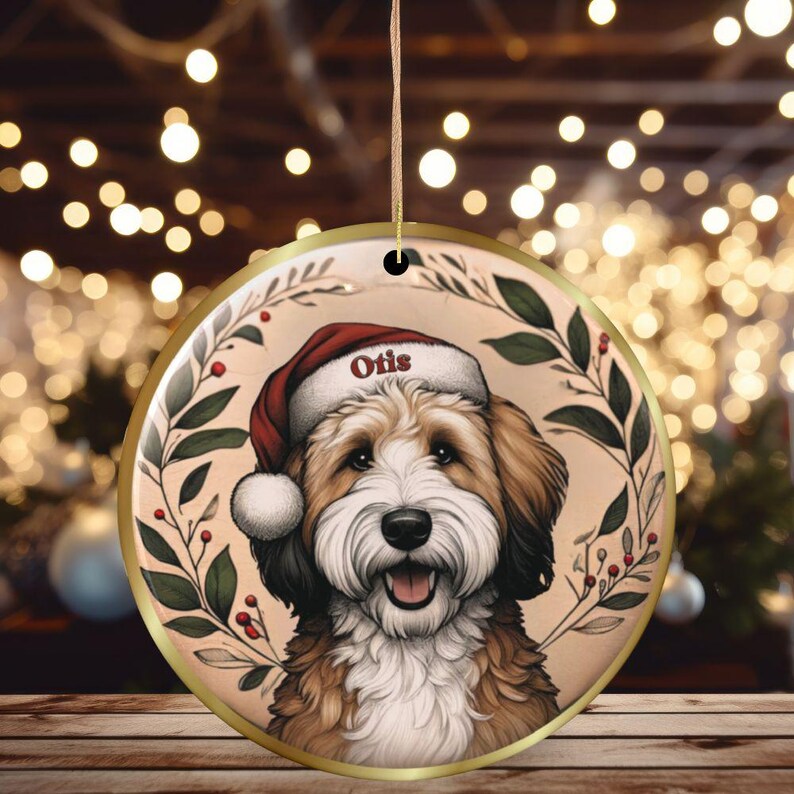 Puede incluir: Un adorno navide&ntilde;o redondo de cer&aacute;mica con borde dorado y cord&oacute;n para colgar. El centro muestra la ilustraci&oacute;n de un perro peludo marr&oacute;n y blanco con un gorro de Pap&aacute; Noel rojo con el nombre Otis en el borde, enmarcado por hojas verdes.