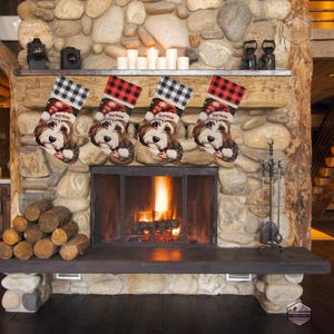 A festive Christmas stocking featuring a whimsical illustrated doodle dog wearing a red Santa hat, surrounded by holiday ornaments and greenery in a warm, detailed Christmas art style. The top of the stocking has a classic red and black buffalo-plaid cuff, and the stocking hangs from a white mantel decorated with evergreen garland and gold ornaments. The stocking design is personalized with your dog’s name, making it a charming and custom holiday décor piece for dog lovers.