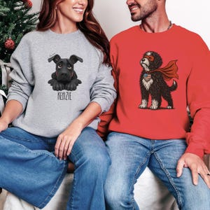 Puede incluir: Dos sudaderas, una gris con un gr&aacute;fico de perro negro y el nombre "KENZIE", la otra roja con un dise&ntilde;o de perro superh&eacute;roe. Ambas son de manga larga y se combinan con vaqueros azules. La imagen muestra un atuendo informal.