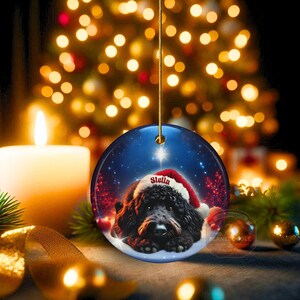 May include: A round Christmas ornament featuring a black dog wearing a Santa hat with the name "Stella". The ornament has a blue background with a star and snow effect. The ornament is hanging from a gold string.