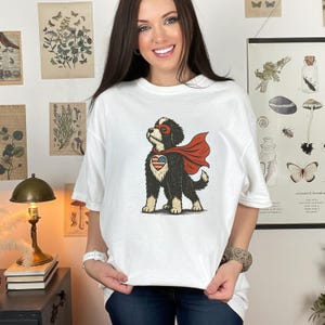 May include: A white short-sleeved t-shirt featuring a graphic illustration of a black and white dog wearing a red superhero cape and a mask. The dog has a heart-shaped patch on its chest with a red, white, and blue American flag pattern.