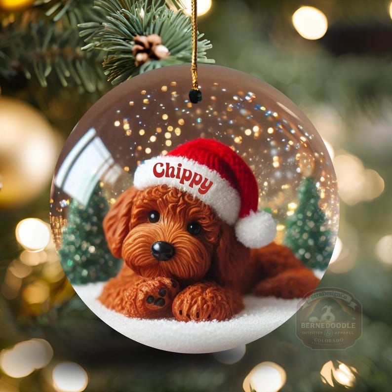 May include: A circular Christmas bauble showing a brown dog wearing a red and white Santa hat. The dog is lying on snow inside a glass globe. The name "Chippy" is written on the hat.