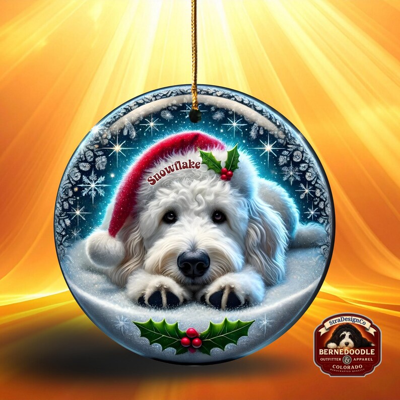 May include: A round Christmas ornament featuring a white dog wearing a red Santa hat with the word "Snowflake". The dog is lying in snow, surrounded by snowflakes. Holly and berries decorate the bottom. A gold string is attached for hanging.