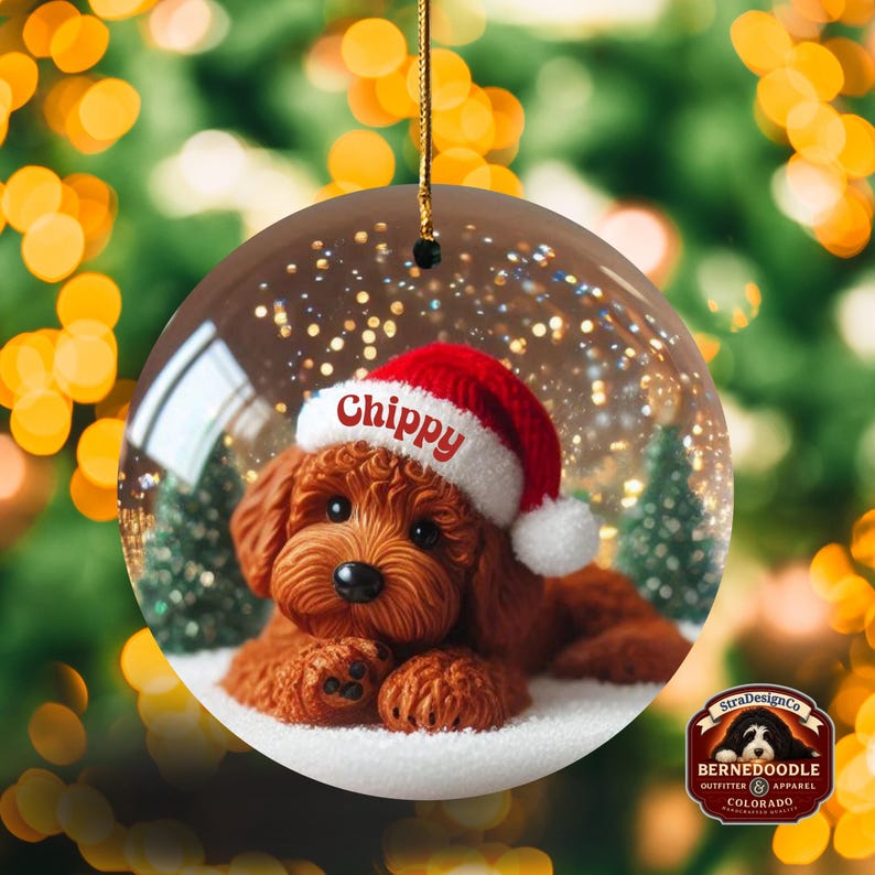 May include: A clear glass Christmas bauble featuring a brown dog wearing a red Santa hat with the name "Chippy" on it. The bauble has a snowy scene with small Christmas trees and a gold string for hanging. The background is blurred with Christmas lights.