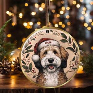 May include: Round Christmas ornament featuring a smiling dog wearing a Santa hat with the name "Otis". The ornament has a gold border and is surrounded by a wreath of green leaves and red berries. The background is blurred with warm, golden lights.