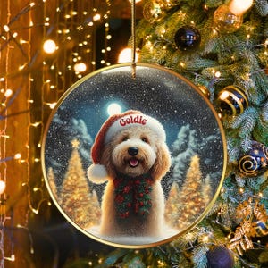 May include: Round Christmas ornament featuring a dog wearing a Santa hat with the name "Goldie" on it. The dog is wearing a red and green garland. The background shows a snowy forest scene with a full moon.