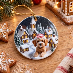 May include: A round ceramic Christmas ornament featuring a golden-brown dog wearing a red and white Santa hat with the name Maggie printed on it. The background shows a snowy village scene with houses, a church, and pine trees.