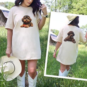 May include: A beige short-sleeve t-shirt featuring a graphic of a brown curly-haired dog on the front chest and a graphic of the same dog from behind on the back. The shirt has a crew neckline and a relaxed fit, showcasing a canine illustration.