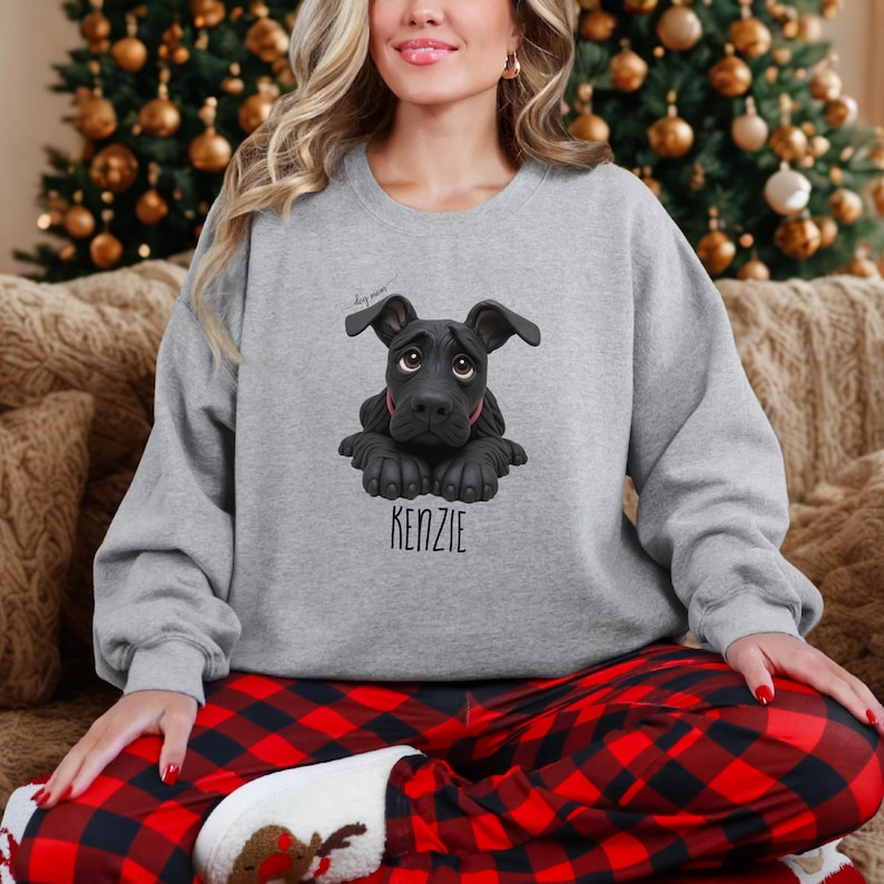 Puede incluir: Sudadera gris claro con un gr&aacute;fico de perro negro y el nombre "KENZIE" impreso debajo. El perro tiene una expresi&oacute;n triste. La sudadera se usa con pantalones de pijama a cuadros rojos y negros.