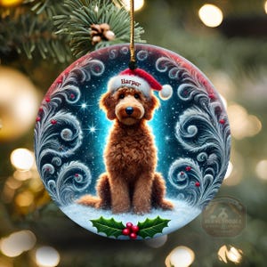 May include: Round Christmas ornament featuring a brown dog wearing a Santa hat with the name "Harper". The ornament has a blue background with white swirls and stars. Holly and berries are at the bottom. A gold string is attached for hanging.
