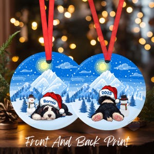 May include: Round Christmas ornament with a winter scene featuring a black and white dog wearing a Santa hat. The ornament has a red ribbon for hanging. The text "Bernie" and "2025" are on the hats. The words "Front And Back Print" are at the bottom.