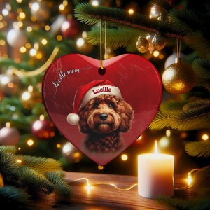 May include: A heart-shaped Christmas ornament with a red background and a portrait of a brown dog wearing a Santa hat. The ornament has the name "Lucille" on the hat and the words "doodle mom" written on the top. The ornament is hanging on a Christmas tree.