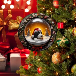 May include: A round Christmas ornament featuring a snow globe design with a black and white dog wearing a Santa hat that says "Bernie". The ornament is surrounded by a wreath of green leaves and red berries. The background shows a Christmas tree and wrapped gifts.
