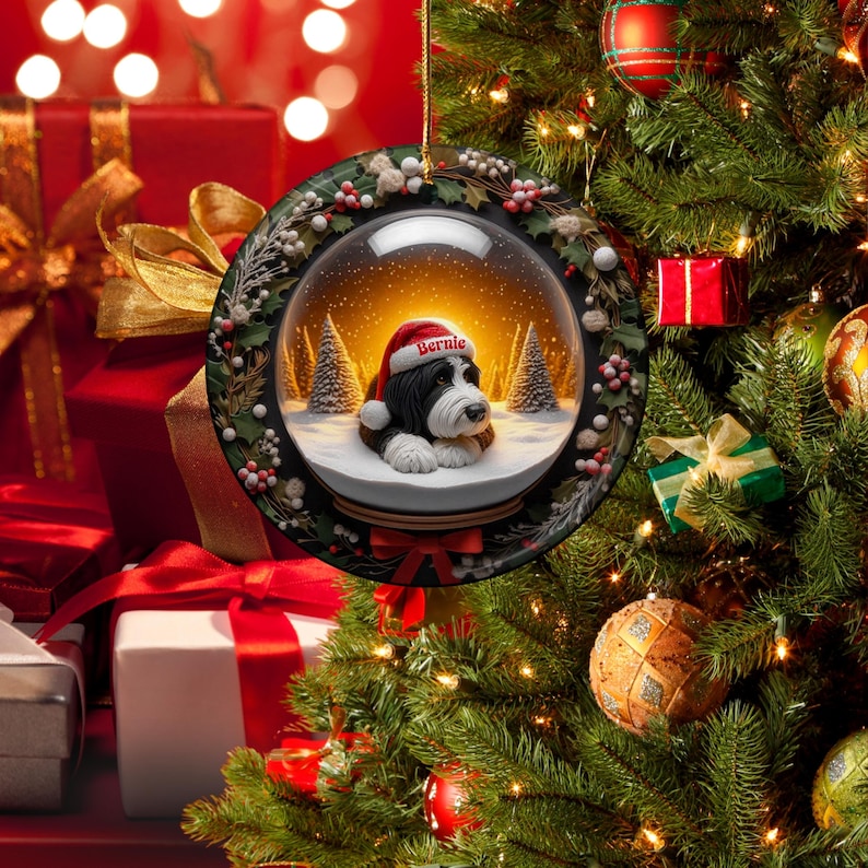 May include: A round Christmas ornament featuring a snow globe design with a black and white dog wearing a Santa hat that says "Bernie". The ornament is surrounded by a wreath of green leaves and red berries. The background shows a Christmas tree and wrapped gifts.