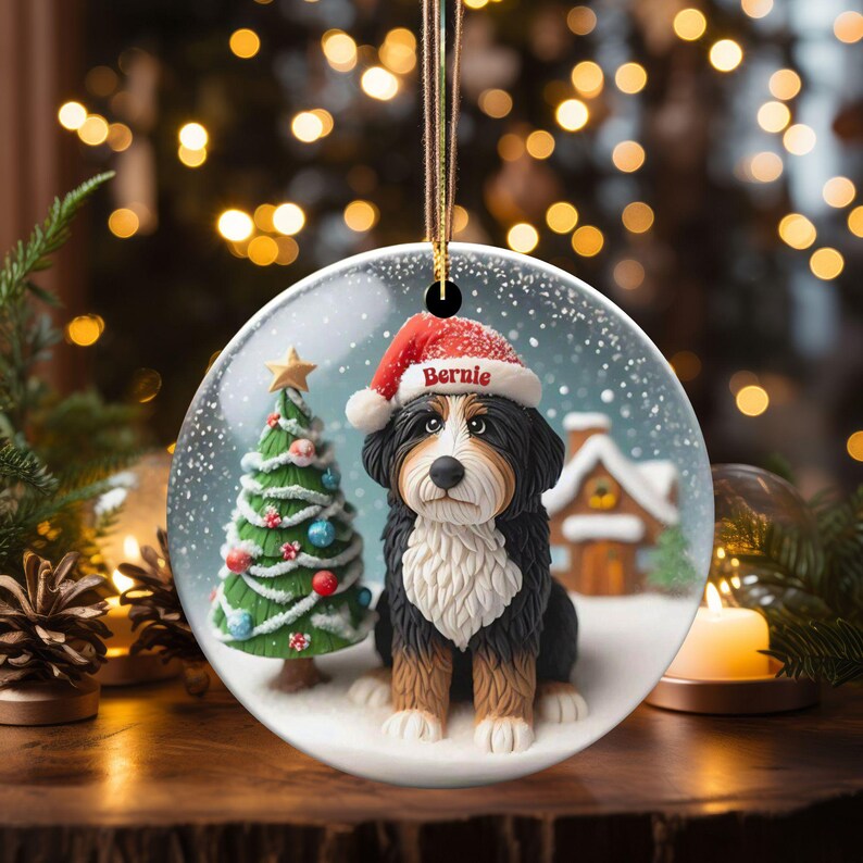 May include: A round Christmas ornament featuring a Bernese Mountain Dog wearing a Santa hat with the name "Bernie". The ornament has a snowy scene with a Christmas tree and a small house. The ornament is hanging by a gold string.