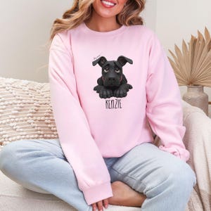 Puede incluir: Sudadera rosa claro con una ilustraci&oacute;n de perro negro y el nombre "KENZIE" en negro. El perro tiene ojos grandes y oscuros y est&aacute; acostado. La sudadera es de color s&oacute;lido.