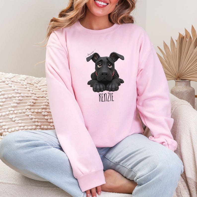 Puede incluir: Sudadera rosa claro con una ilustraci&oacute;n de perro negro y el nombre "KENZIE" en negro. El perro tiene ojos grandes y oscuros y est&aacute; acostado. La sudadera es de color s&oacute;lido.