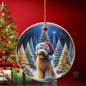 May include: A round Christmas ornament with a gold trim, featuring a fluffy, light brown dog wearing a Santa hat with the name "Goldie" on it. The background shows a snowy forest scene with Christmas trees. A Christmas tree and presents are in the background.