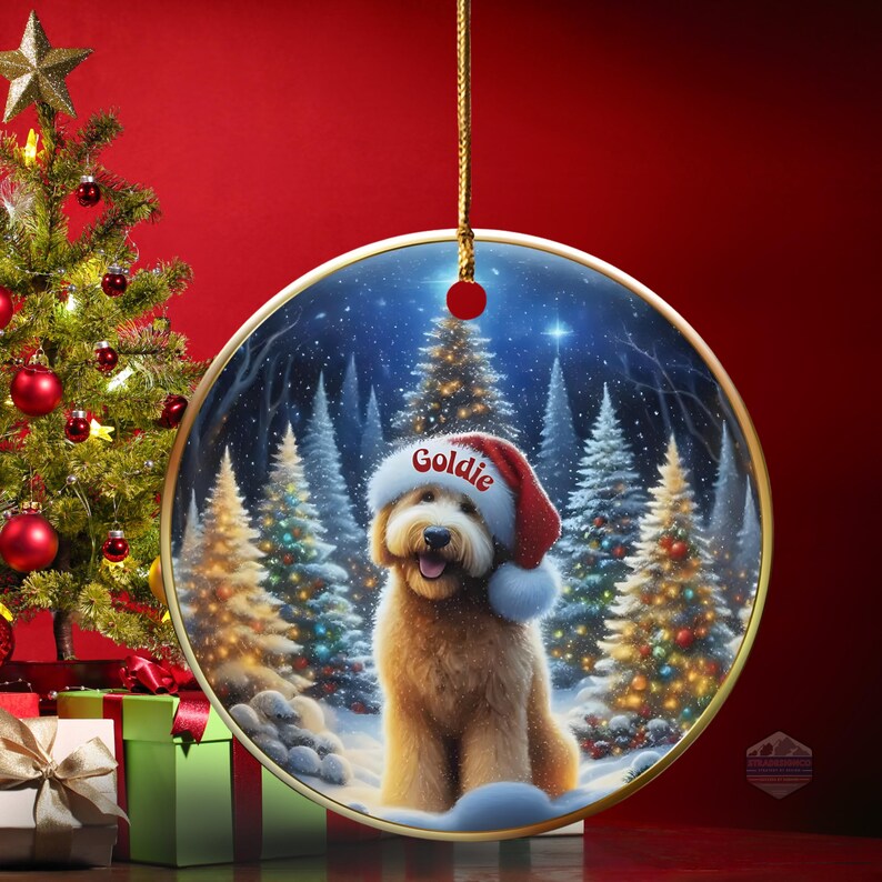 May include: A round Christmas ornament with a gold trim, featuring a fluffy, light brown dog wearing a Santa hat with the name "Goldie" on it. The background shows a snowy forest scene with Christmas trees. A Christmas tree and presents are in the background.