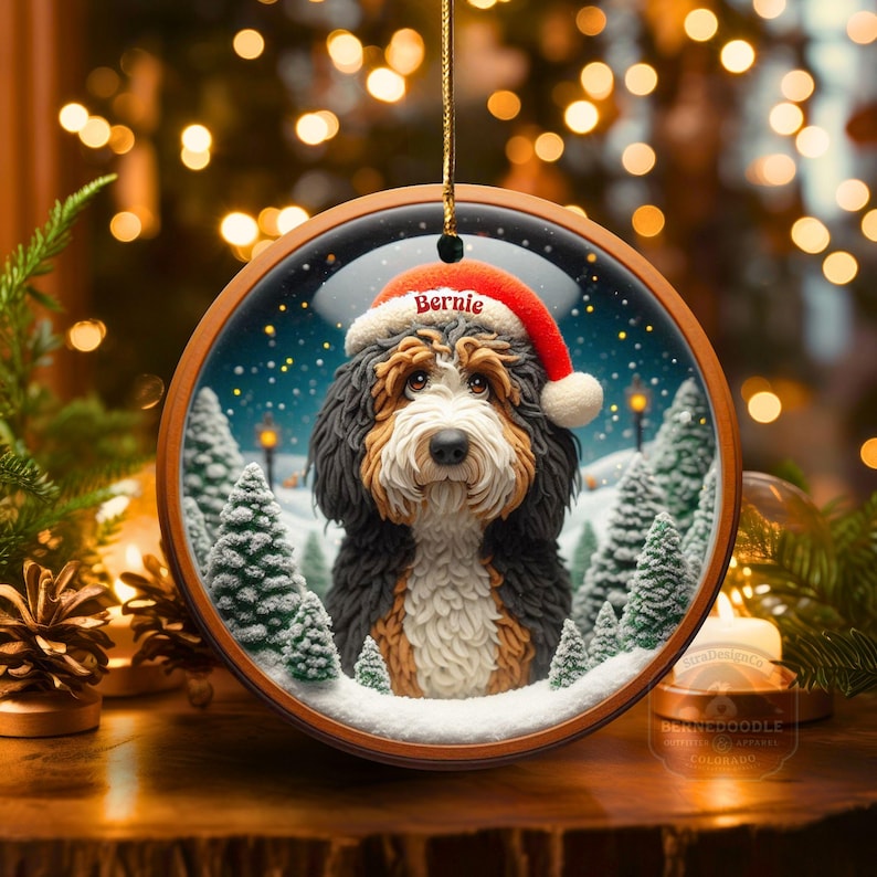 Op de afbeelding: Een ronde houten kerstversiering met een Bernedoodle hond met een kerstmuts met de naam "Bernie". De versiering heeft een winterse achtergrond met besneeuwde bomen. Het hangt aan een gouden touwtje.