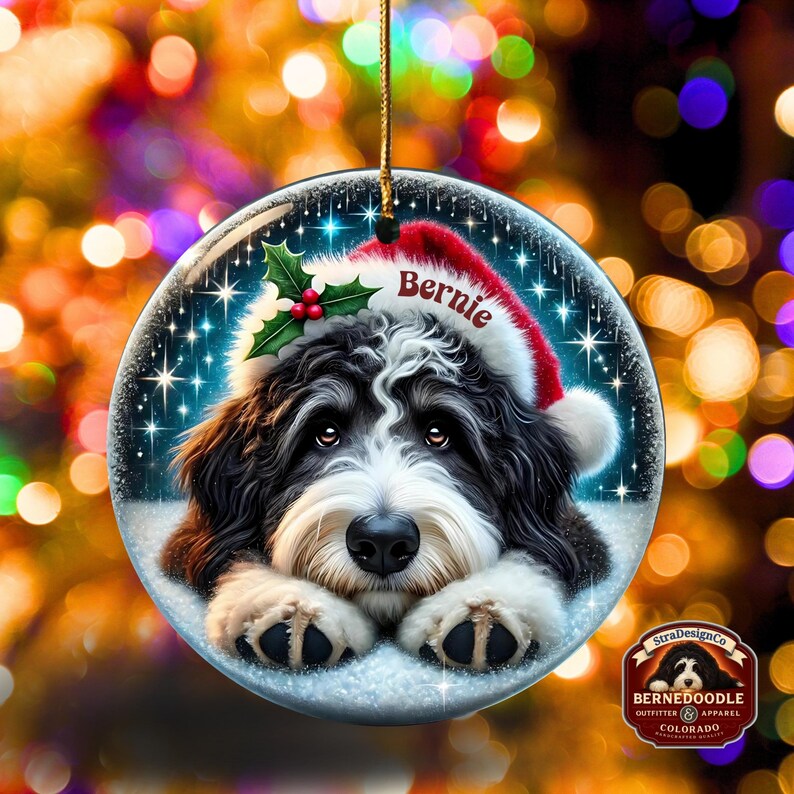 May include: A round Christmas ornament featuring a Bernedoodle dog wearing a Santa hat with the name "Bernie." The ornament has a snowy background with stars and holly. A gold string is attached for hanging.