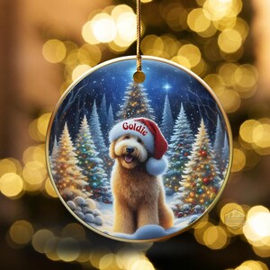 May include: Round Christmas ornament featuring a golden doodle dog wearing a Santa hat with the name "Goldie". The background shows a snowy forest with Christmas trees. The ornament is hanging with a gold string.