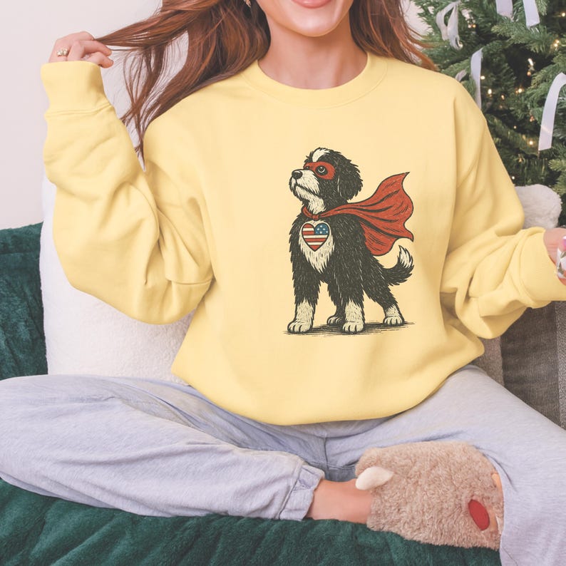 May include: A light yellow sweatshirt featuring a cartoon dog dressed as a superhero. The dog has a red cape, a mask, and a heart-shaped emblem with an American flag design on its chest. The sweatshirt is a solid color.