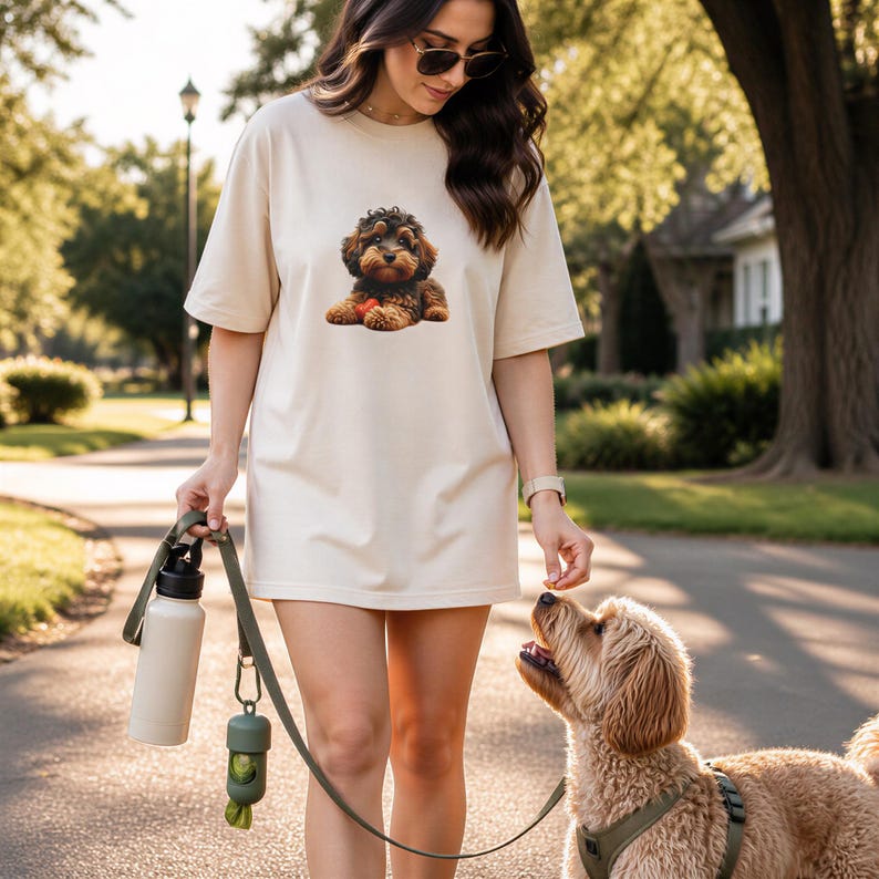 A woman in a white t-shirt with a dog on it is walking her dog on a leash in a park.