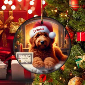 Pu&ograve; includere: Un ornamento natalizio rotondo con un cane di tipo golden retriever che indossa un cappello da Babbo Natale con il nome "Frannie". L'ornamento ha un cordino dorato per appenderlo e include un certificato "Good Dog". Lo sfondo mostra un camino e un albero di Natale.