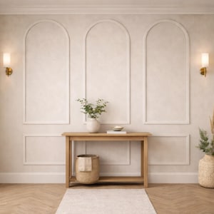 Op de afbeelding: Een neutraal interieur met een houten console, een rieten mand en een wit tapijt. De muren zijn versierd met gewelfde panelen en wandkandelaars. Decoratieve vazen en groen voegen een natuurlijk tintje toe. De vloer heeft een visgraatpatroon.