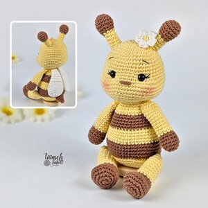 May include: A handmade crochet bee doll with a yellow and brown striped body, brown accents, and white wings. The bee has a smiling face, a white flower on its head, and is sitting on a wooden base. The doll is a charming, whimsical creation.