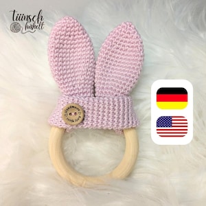Puede incluir: Un mordedor para bebé tejido a mano, de color rosa claro, con forma de conejito y grandes orejas. El mordedor tiene un anillo de madera y un botón de madera con la inscripción "handmade with love". El logotipo "Tiinsch häkelt" es visible.