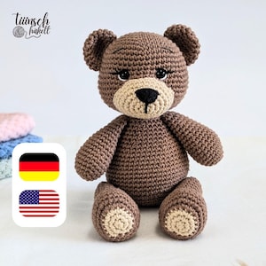 Könnte beinhalten: Ein handgefertigter brauner Häkelteddybär mit beiger Schnauze und Pfoten. Der Bär hat eine schwarze Nase und kleine, dunkle Augen. Der Text "Tiinisch Hakell" befindet sich in der oberen linken Ecke. Der Bär sitzt auf einer weißen Oberfläche.