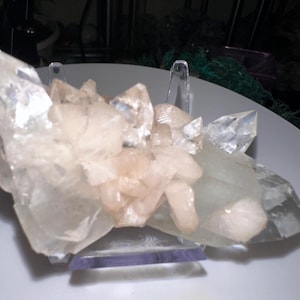 May include: A cluster of clear and peach-colored crystals, displayed on a clear plastic stand. The crystals have sharp, pointed edges and a translucent appearance. The arrangement suggests a natural, geological specimen.