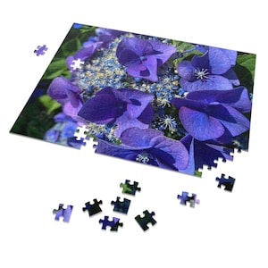 May include: A partially assembled jigsaw puzzle depicting a close-up of vibrant purple hydrangea flowers. Scattered puzzle pieces surround the edges, with some still connected. The image highlights the intricate details of the floral arrangement.