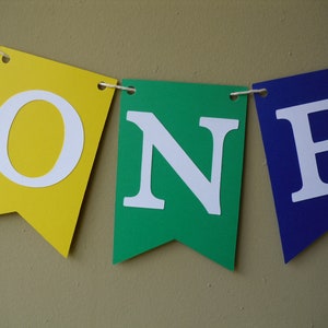 May include: A colorful banner with the word "ONE" in white letters on a yellow, green, and blue background. The banner is perfect for a first birthday party.