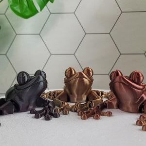 May include: Three 3D printed frog figurines in different metallic finishes: black, gold, and copper. The frogs are posed with their legs outstretched and their eyes closed.