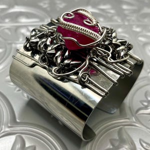 May include: A silver cuff bracelet with a pink gemstone wrapped in silver wire. The gemstone is surrounded by silver wire designs.