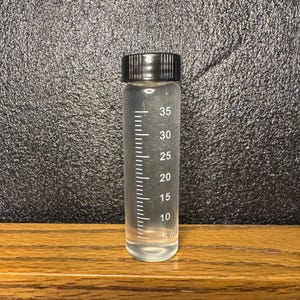 Può includere: Un flacone cilindrico in plastica trasparente con tappo a vite nero. Il flacone presenta tacche di misurazione stampate in bianco che vanno da 5 ml a 35 ml lungo il lato, indicando la sua capacità di contenere liquidi o piccoli oggetti.