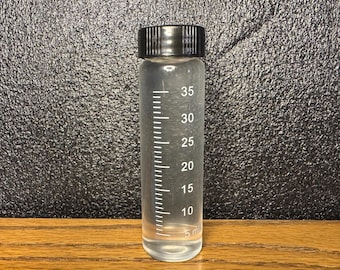 May include: A clear cylindrical plastic vial with a black screw-top lid. The vial features white printed measurement markings ranging from 5 ml to 35 ml along the side, indicating its capacity for holding liquid or small items in a laboratory setting.