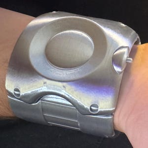 May include: A wide, brushed silver-tone metal cuff bracelet featuring a prominent circular center indentation and a smooth, curved surface. The design includes visible screw-like details at the corners and a segmented band structure that wraps around.