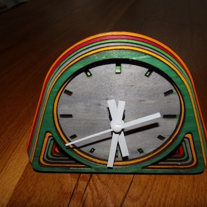 Peut inclure: Une horloge en bois colorée avec un cadran et des aiguilles blancs. L'horloge a un design en couches avec des cercles concentriques de vert, rouge et orange.