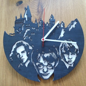 Puede incluir: Un reloj de madera azul oscuro cortado con láser con las siluetas de tres personajes y un castillo. Las manecillas del reloj son blancas con un centro rojo. Un artículo decorativo único.