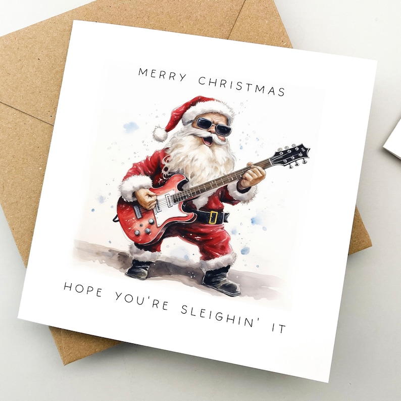 May include: A Christmas card featuring Santa Claus playing a red electric guitar while wearing sunglasses. The card has the text "MERRY CHRISTMAS" at the top and "HOPE YOU'RE SLEIGHIN' IT" at the bottom. The card is on a white background with a brown envelope.