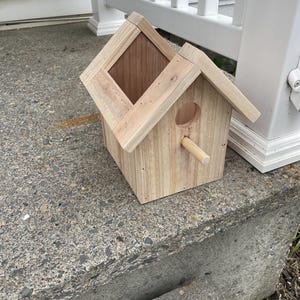May include: A small, wooden birdhouse with a pitched roof and a round entrance hole. The birdhouse is constructed from light-colored wood and features a wooden perch extending from the front. The back of the birdhouse has a diamond-shaped opening.