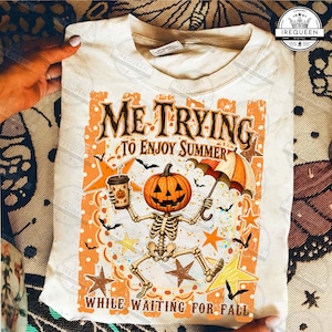 Pode incluir: Uma t-shirt cor de creme com um gráfico de um esqueleto com cabeça de abóbora segurando um copo de café e um guarda-chuva. O texto "ME TRYING TO ENJOY SUMMER WHILE WAITING FOR FALL" está rodeado por estrelas laranja, morcegos e salpicos.