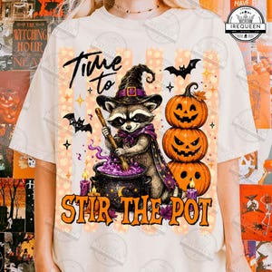 May include: A cream-colored t-shirt featuring a graphic of a raccoon wearing a witch hat and cape, stirring a purple potion in a black cauldron. Beside the raccoon are three stacked jack-o'-lanterns, bats, and the text Time to STIR THE POT.