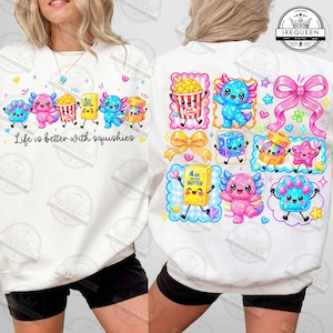 May include: A white sweatshirt featuring a colorful graphic print of various cartoon characters and objects, including axolotls, popcorn, butter, and bows. The front displays the text Life is better with squishies in black script lettering.